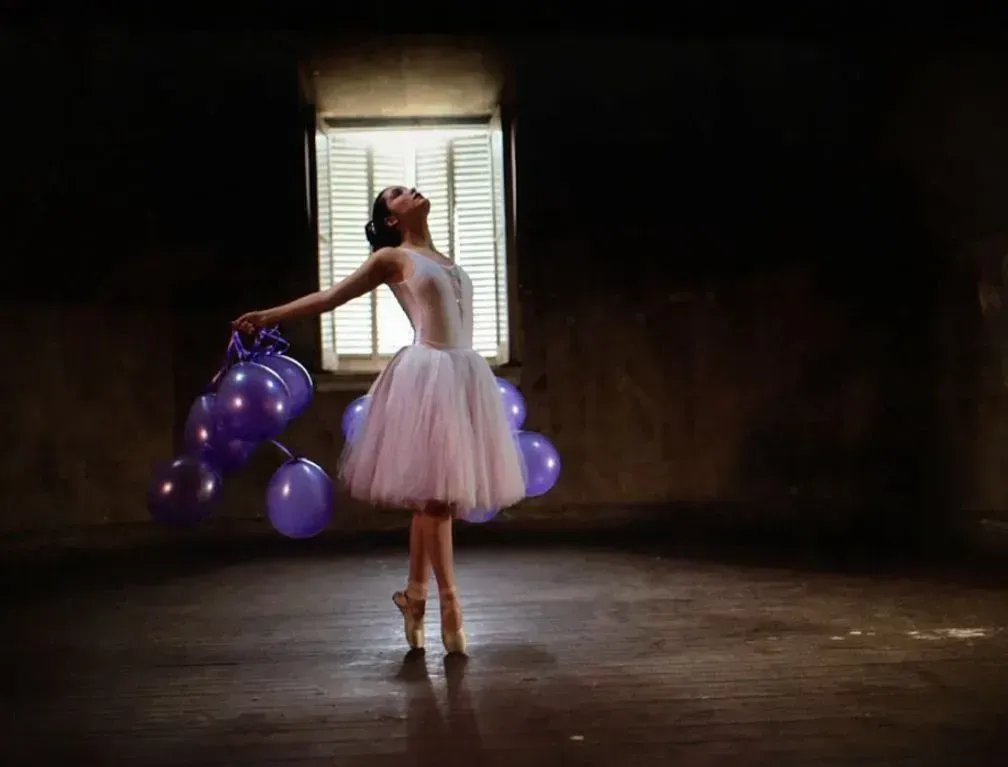 Una bailarina con un tutú blanco posa con globos morados en una habitación con poca luz junto a una ventana luminosa.