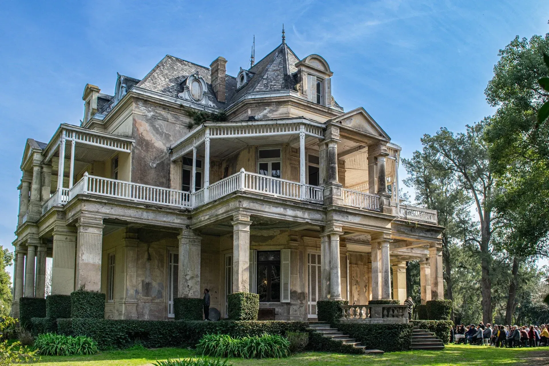 Mansión victoriana con porche que rodea la casa y barandillas blancas, situada en un césped verde bajo un cielo azul.