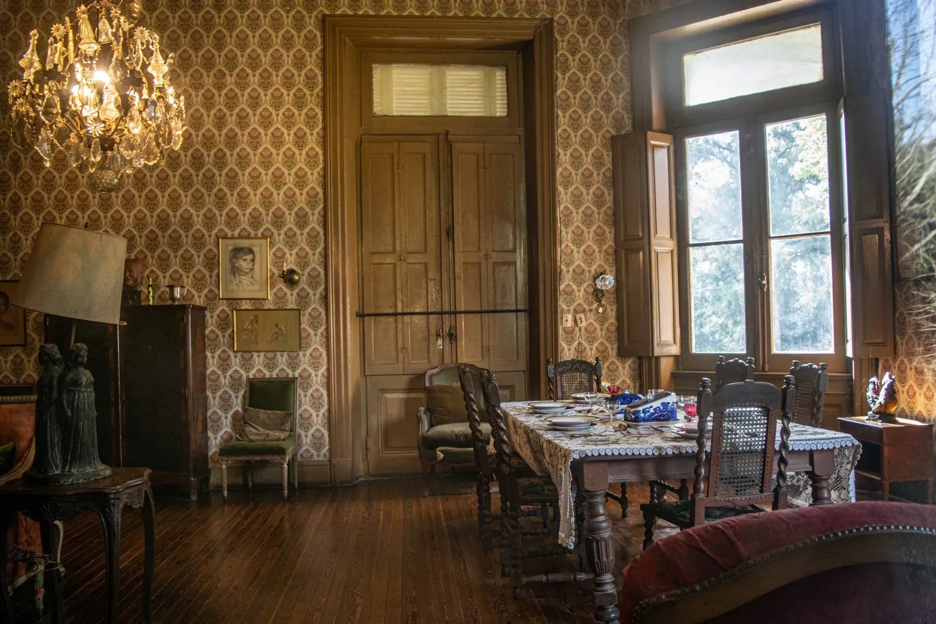 Comedor antiguo con iluminación tenue, papel tapiz estampado, lámpara de araña, ventanas altas y una mesa puesta.