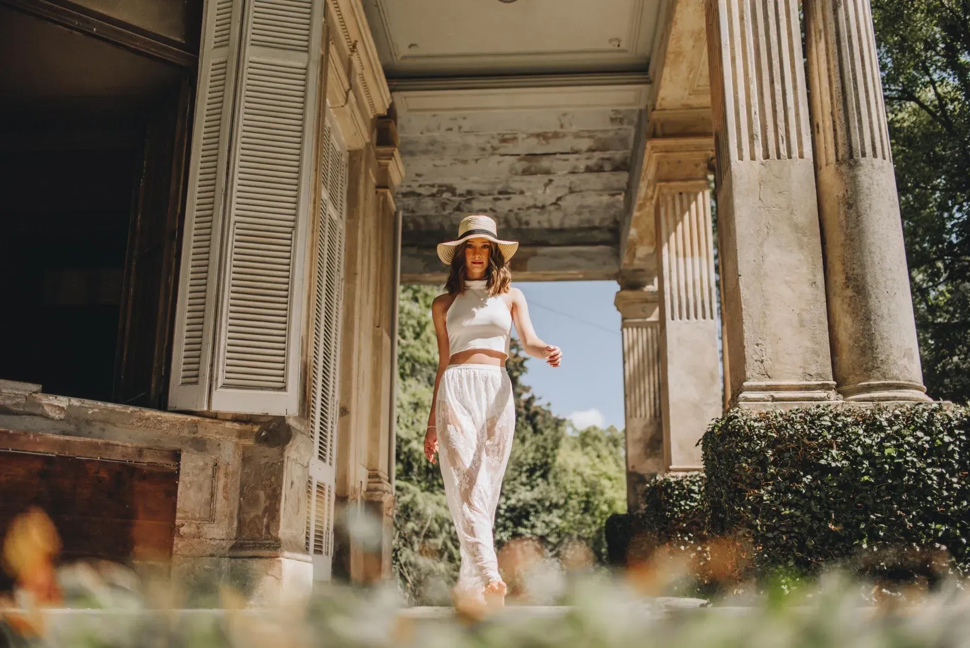 Mujer con vestido y sombrero blancos caminando por un porche soleado con columnas y contraventanas.