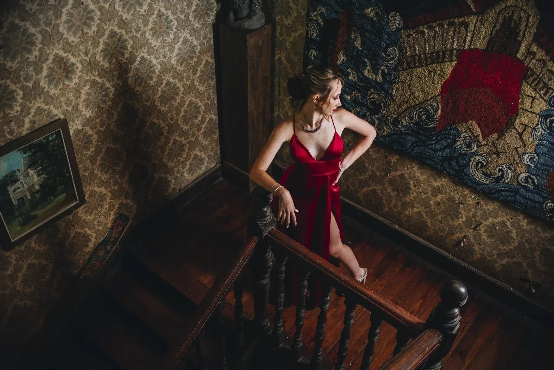 Mujer con vestido rojo posando en una escalera en una habitación con poca luz y papel tapiz estampado.