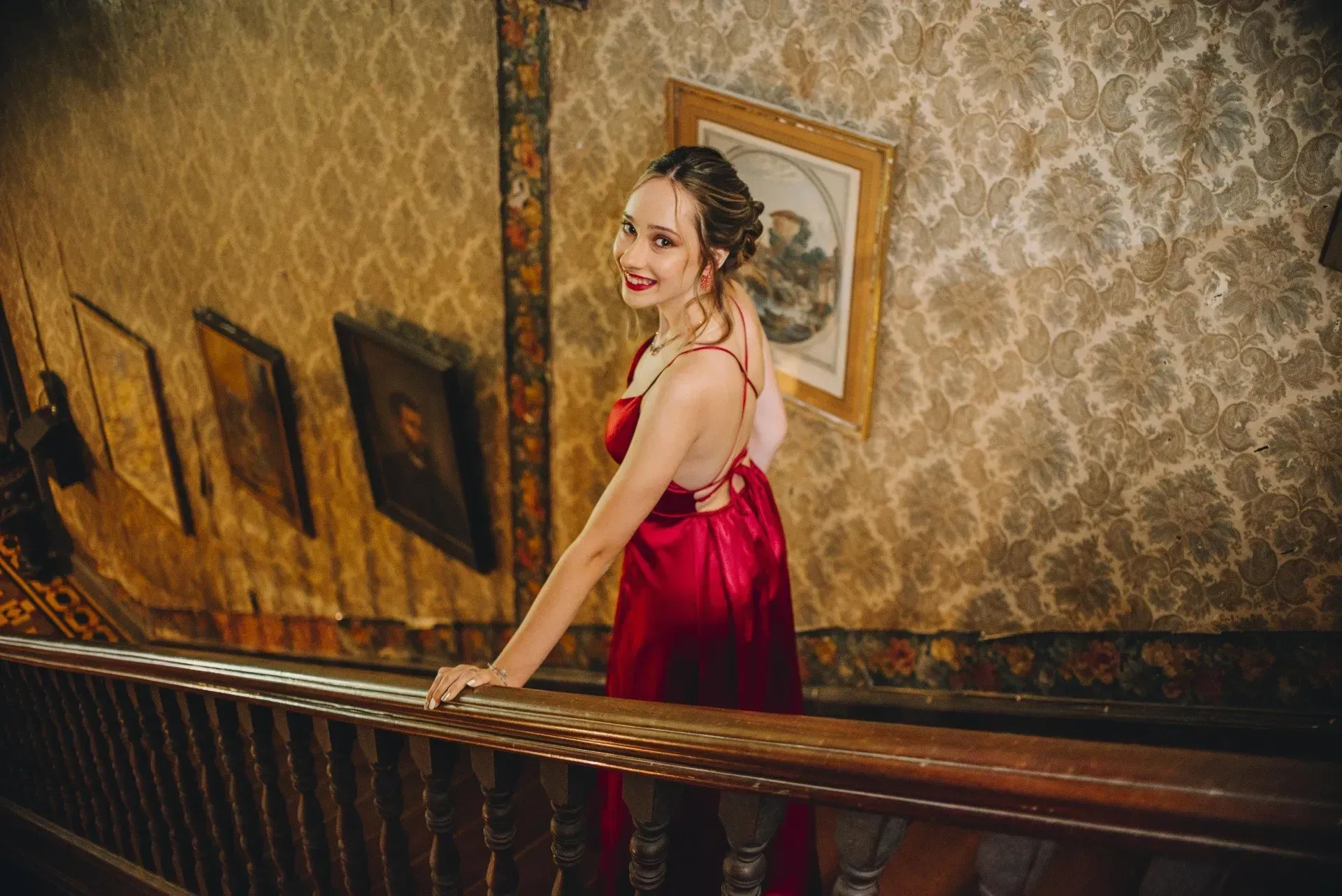 Mujer con vestido rojo en una escalera, mirando hacia atrás con una sonrisa en un pasillo de estilo antiguo.