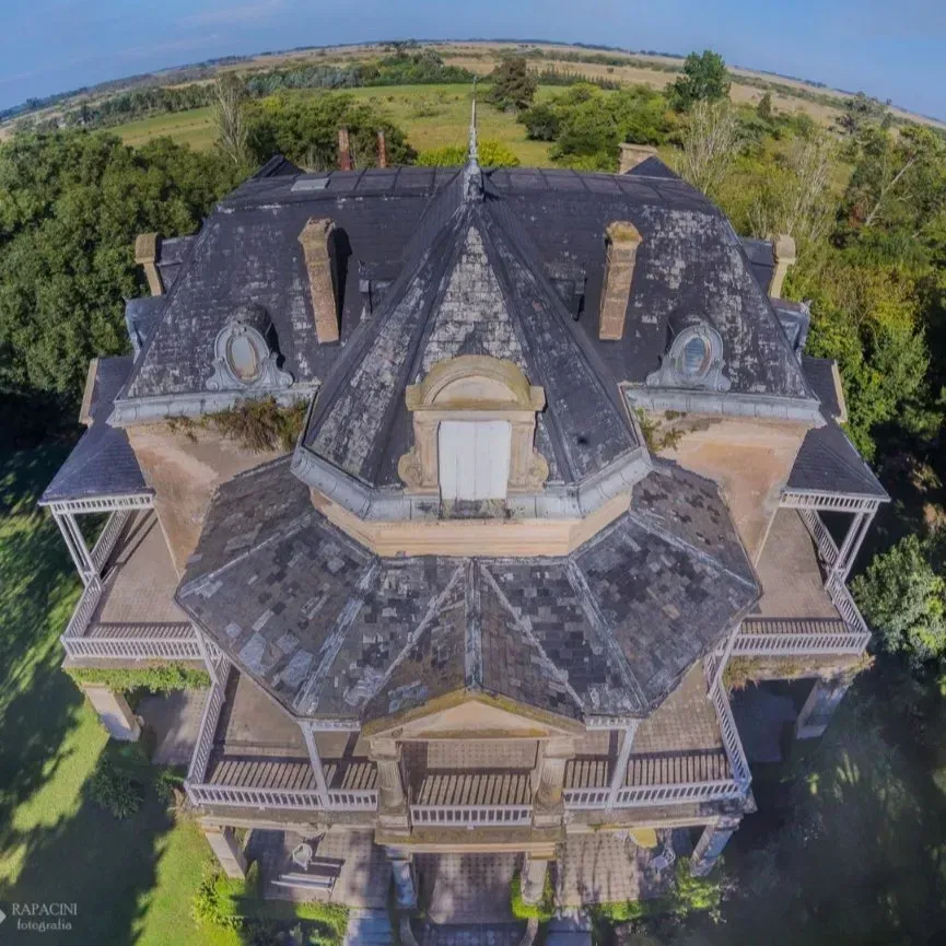 Vista aérea de una gran casa histórica con techo oscuro, rodeada de árboles y paisaje abierto.