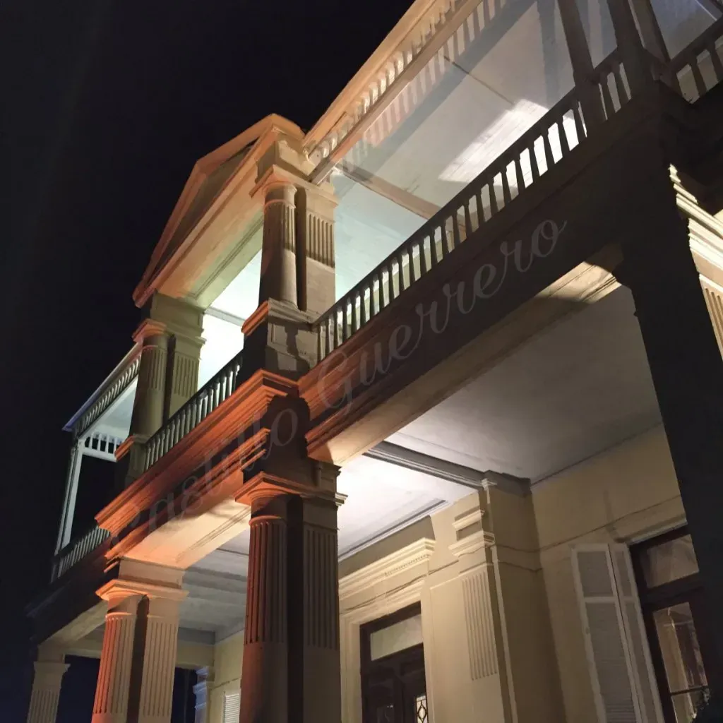 Porche iluminado de una casa color crema por la noche, con columnas y barandilla del balcón contra el cielo oscuro.
