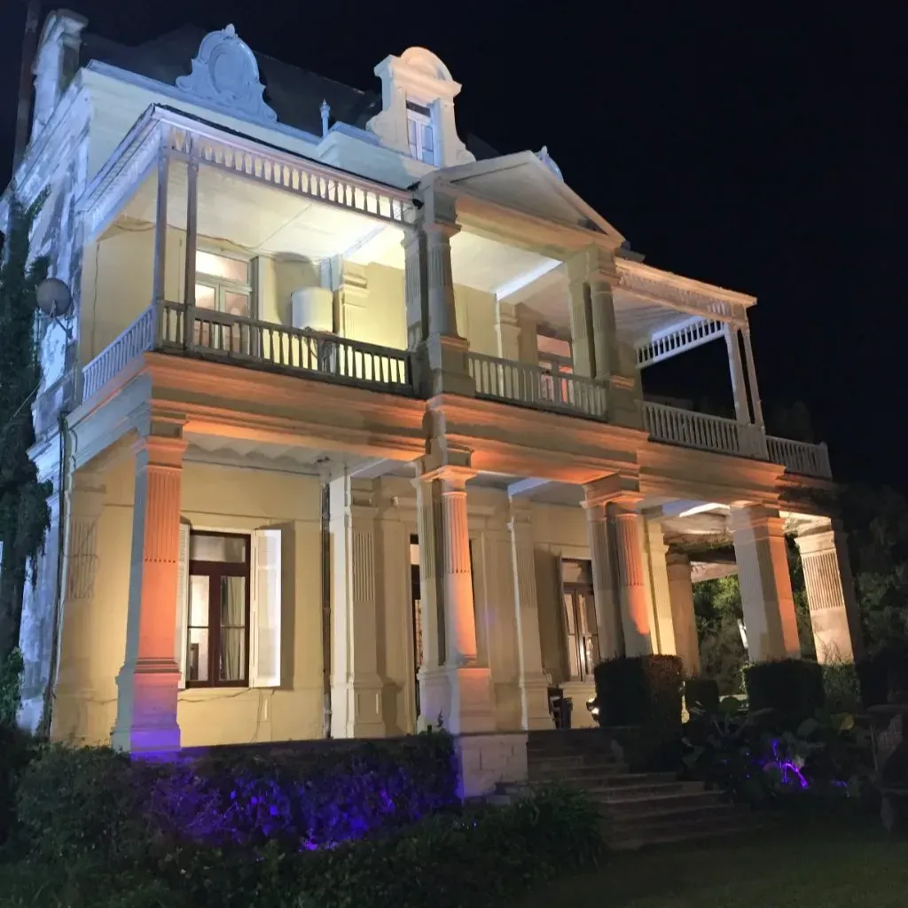 Mansión histórica iluminada por la noche con columnas, balcones y jardines iluminados con luces violetas.