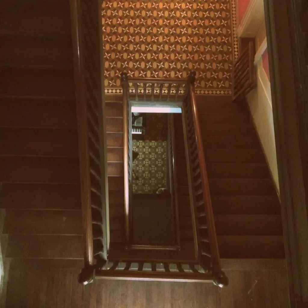 Vista desde arriba de una escalera de madera con papel pintado estampado y un rellano con espejo debajo.