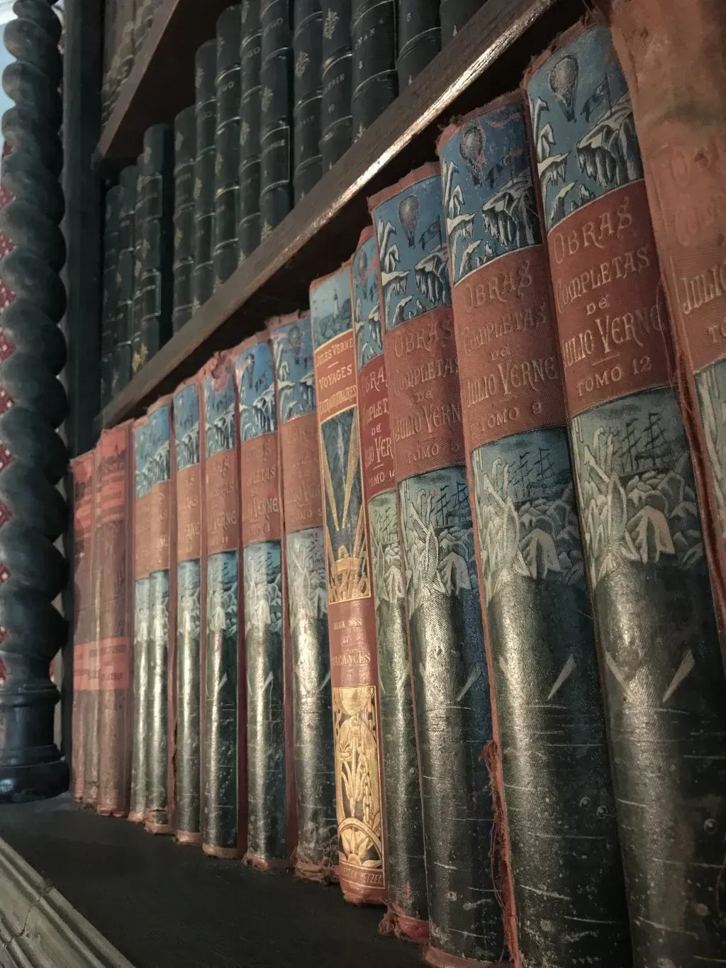 Hilera de libros ornamentados, encuadernados en cuero rojo y azul, en una estantería oscura.