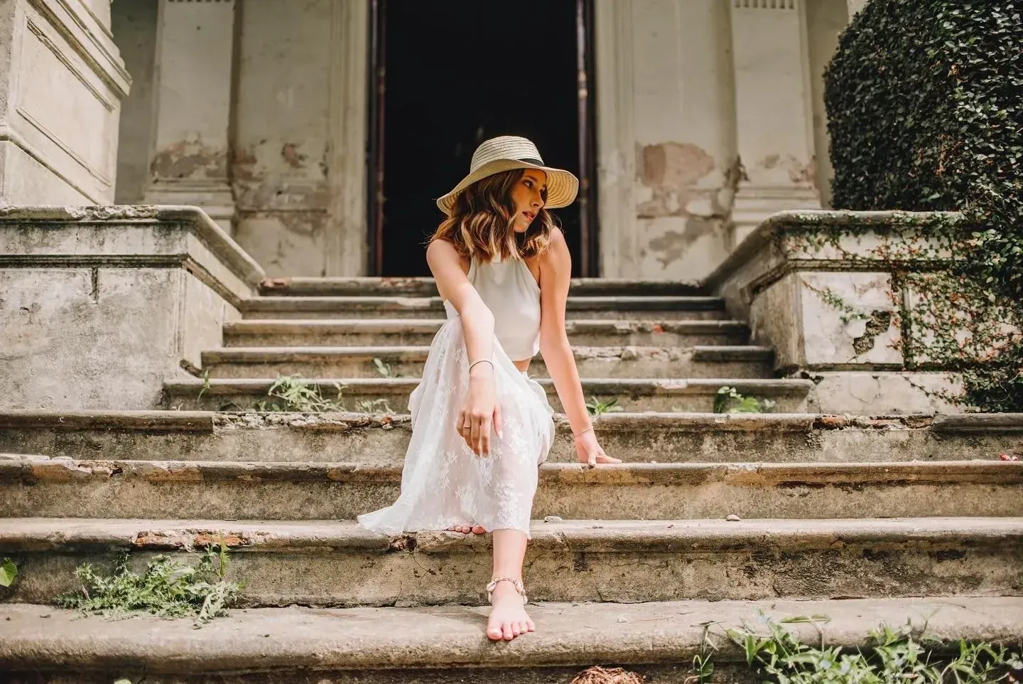 Mujer con vestido blanco y sombrero de paja bajando escalones de piedra en el exterior de un edificio.