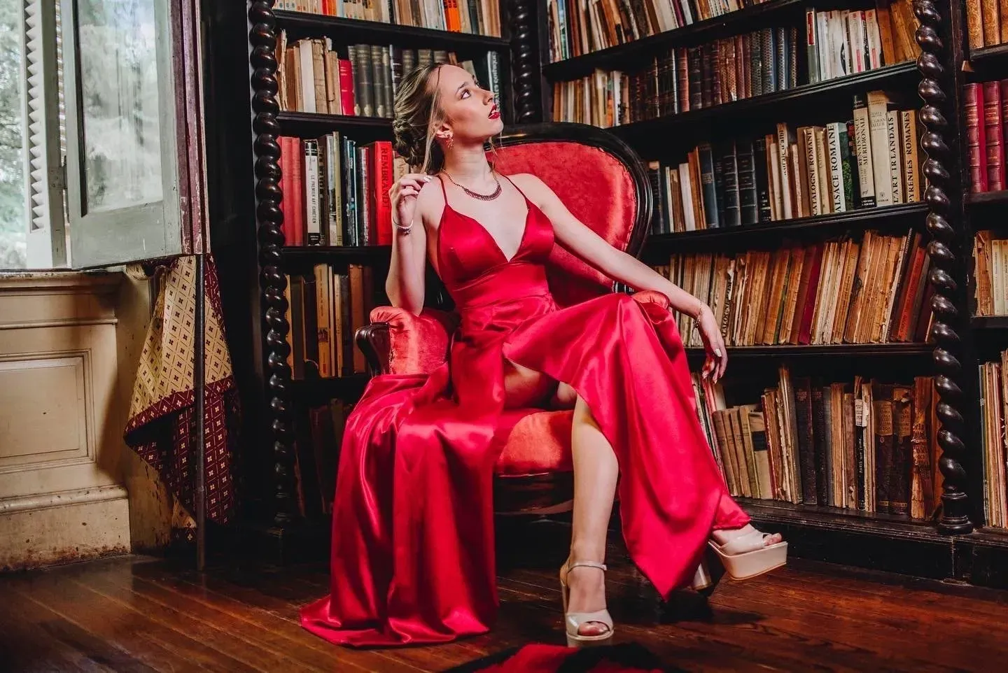 Mujer con un vestido de satén rojo sentada en una biblioteca, rodeada de estanterías.