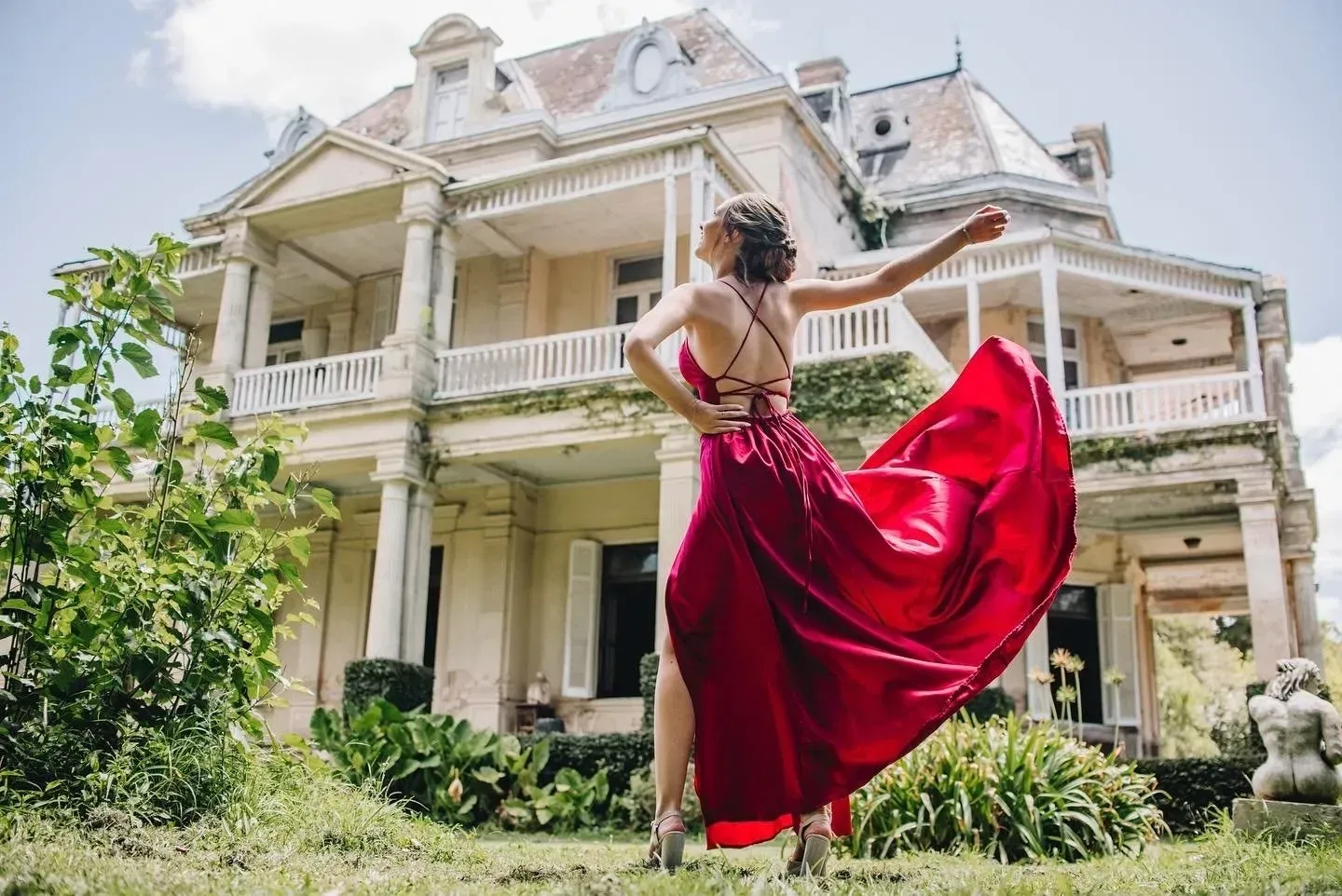Mujer con un vaporoso vestido rojo posando frente a una imponente mansión victoriana blanca con un porche que la rodea.