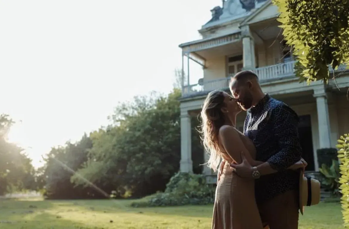 Una pareja se abraza en el césped frente a una casa histórica bañada por el sol.