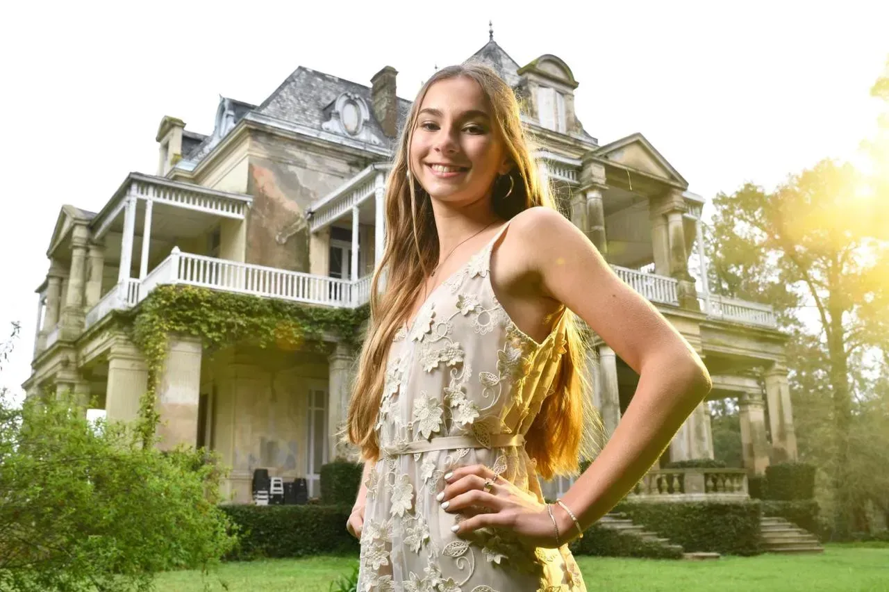 Mujer sonriente con un vestido beige de pie frente a una gran casa histórica con jardín bajo la brillante luz del sol.
