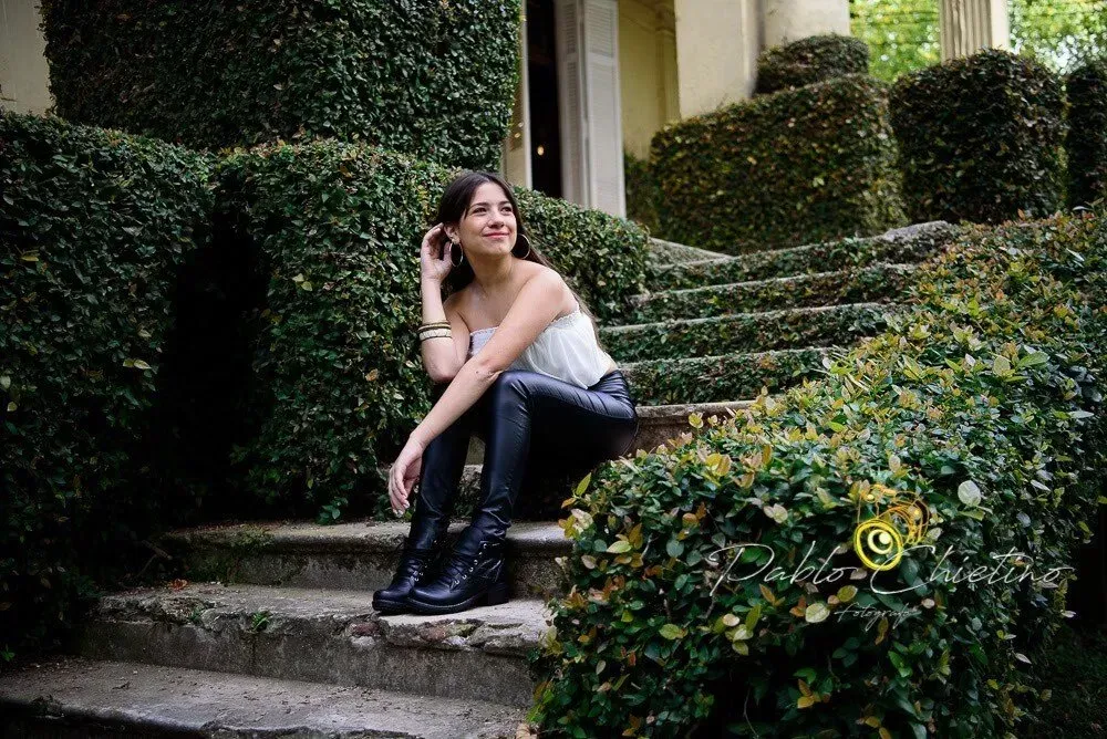 Mujer sentada en escalones de piedra en un jardín, sonriendo junto a setos recortados y vegetación.