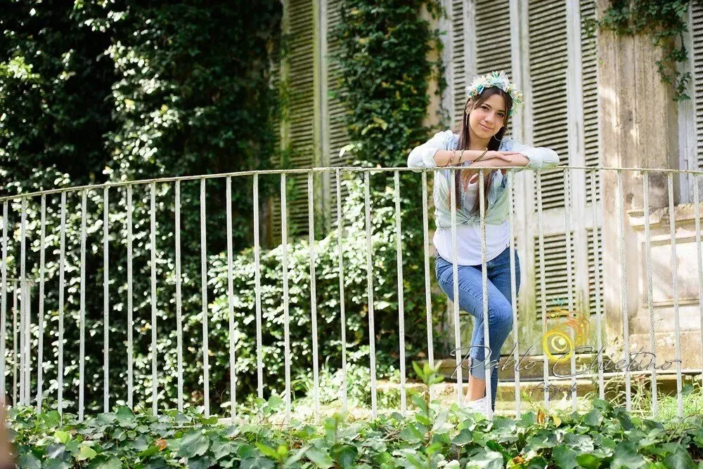 Mujer apoyada en una valla blanca en un jardín exuberante, vestida con vaqueros y una blusa clara.