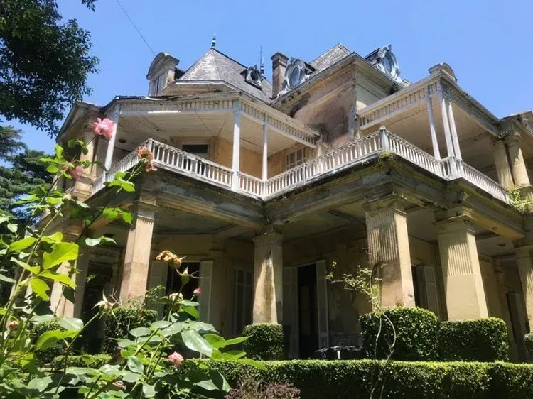 Casa victoriana con porche que rodea la casa, molduras blancas y un exuberante jardín verde bajo un cielo azul.