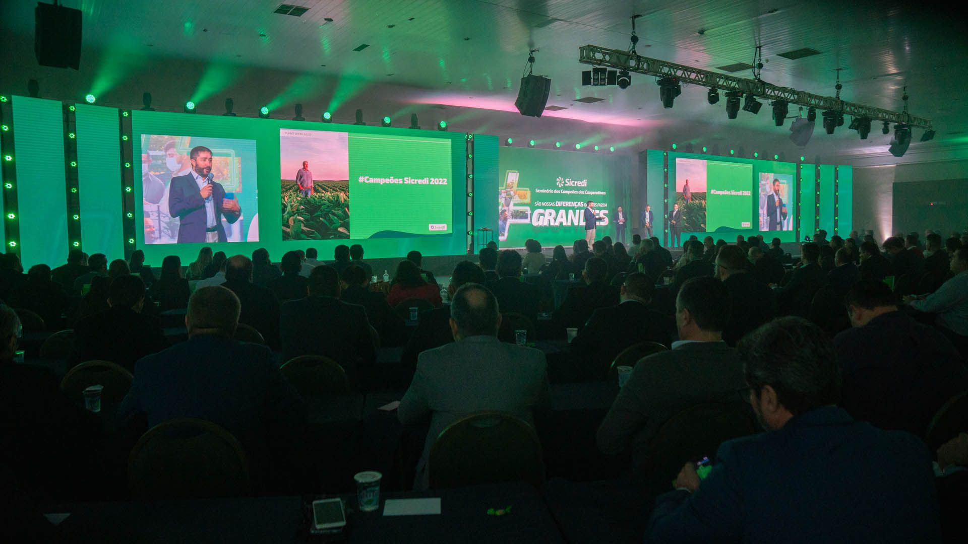 Público de uma conferência de frente para um palco iluminado de verde com telões e palestrantes em um evento ao vivo.