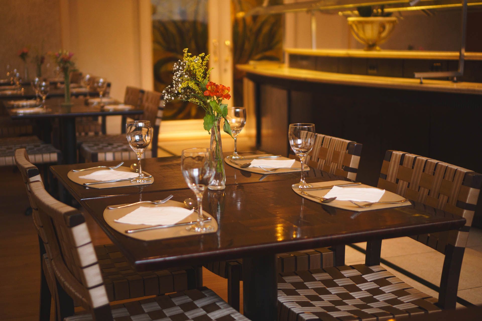 Mesa de restaurante vazia, posta com pratos, copos, guardanapos e um arranjo floral no centro, sob iluminação aconchegante.