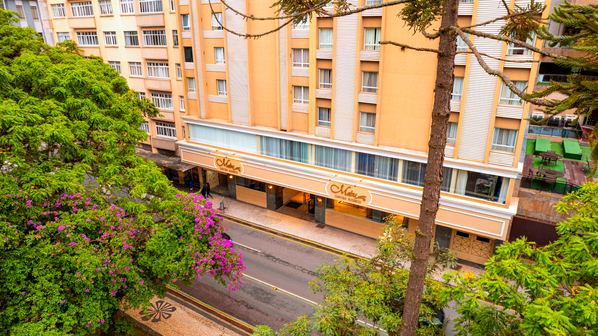 Prédio de apartamentos com varandas laranja ao lado de uma rua arborizada e arbustos floridos cor-de-rosa.