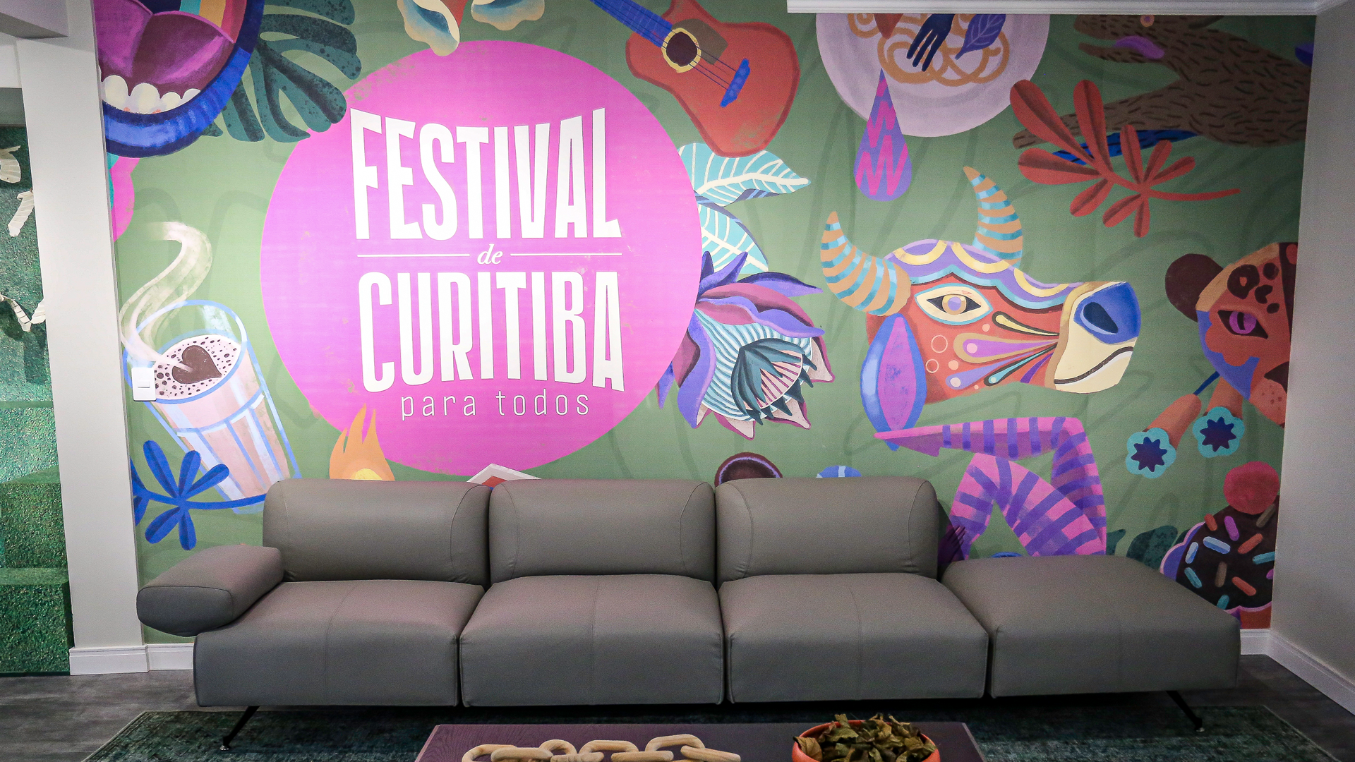Mural colorido do Festival Curitiba atrás de um sofá cinza em uma área de estar moderna.