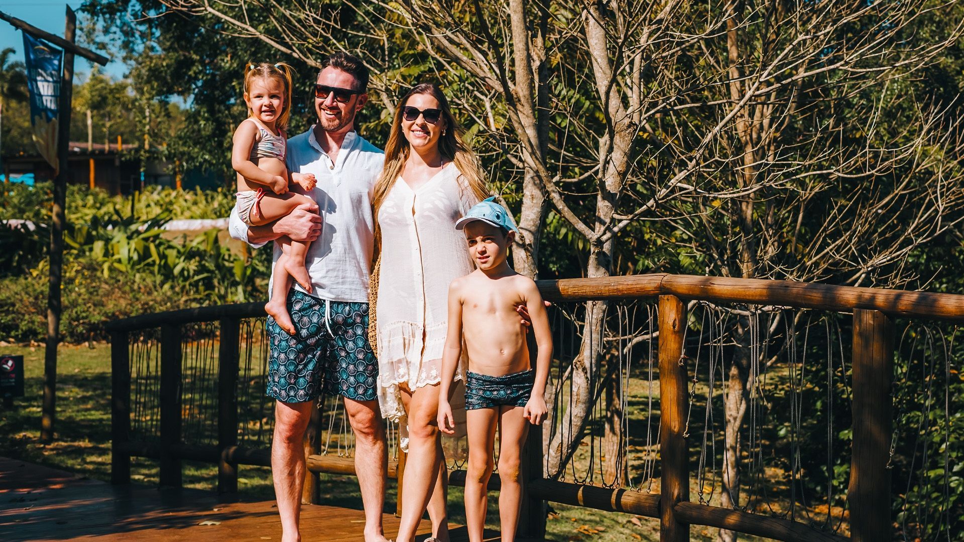 Família em pé numa ponte de madeira numa área arborizada e ensolarada, sorrindo para a câmera.