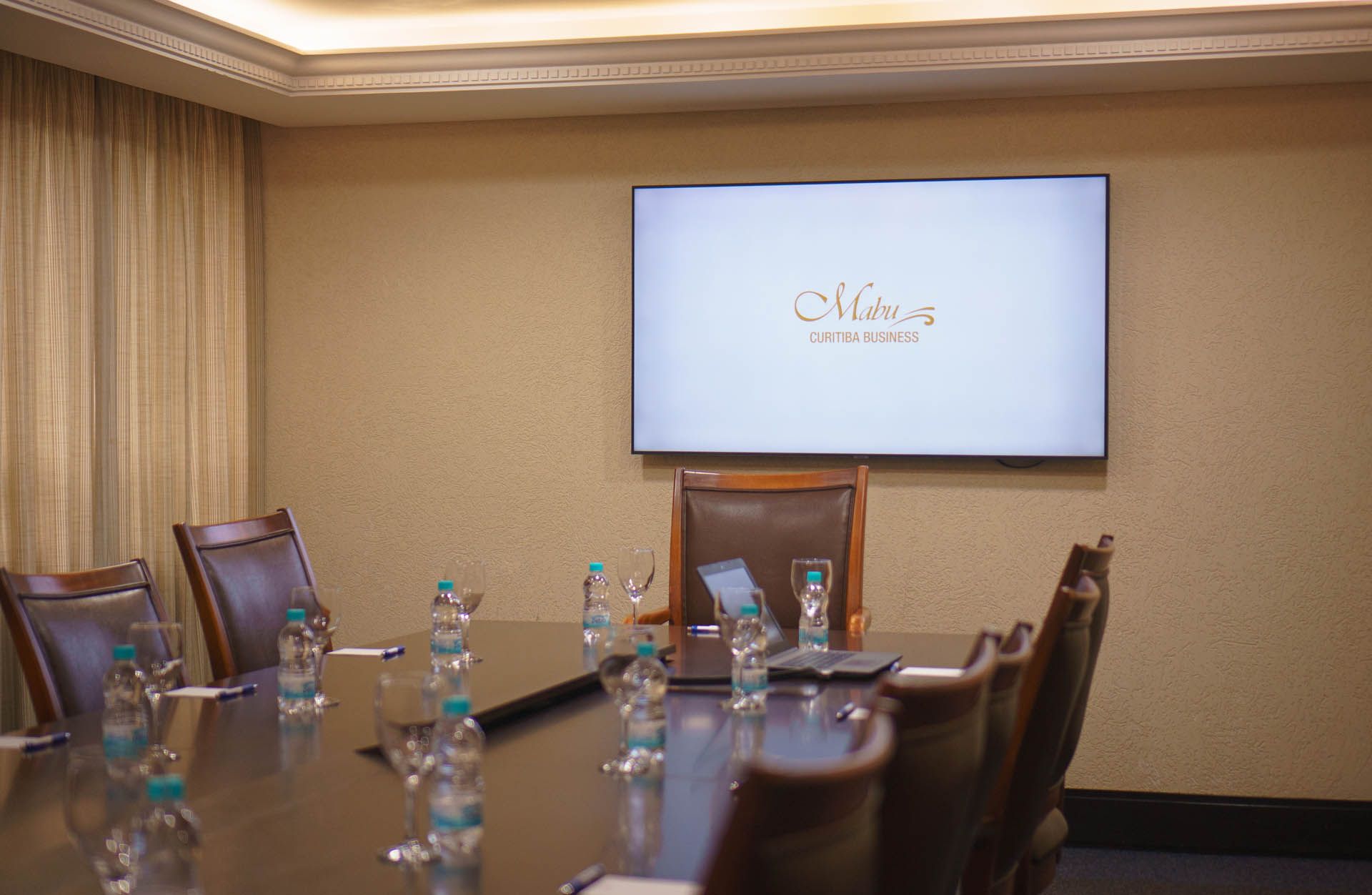 Sala de conferências vazia com uma mesa, cadeiras, garrafas de água e uma tela fixada na parede exibindo um logotipo.