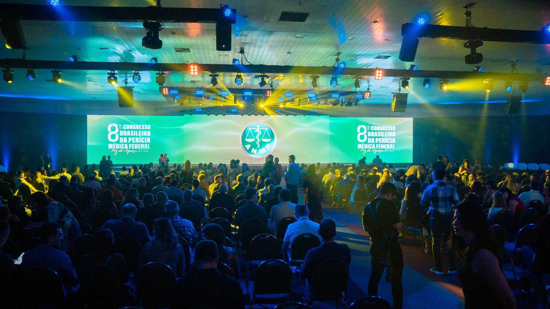 Sala de conferências lotada com palco iluminado, telas azul-esverdeadas e silhuetas da plateia voltadas para os apresentadores.