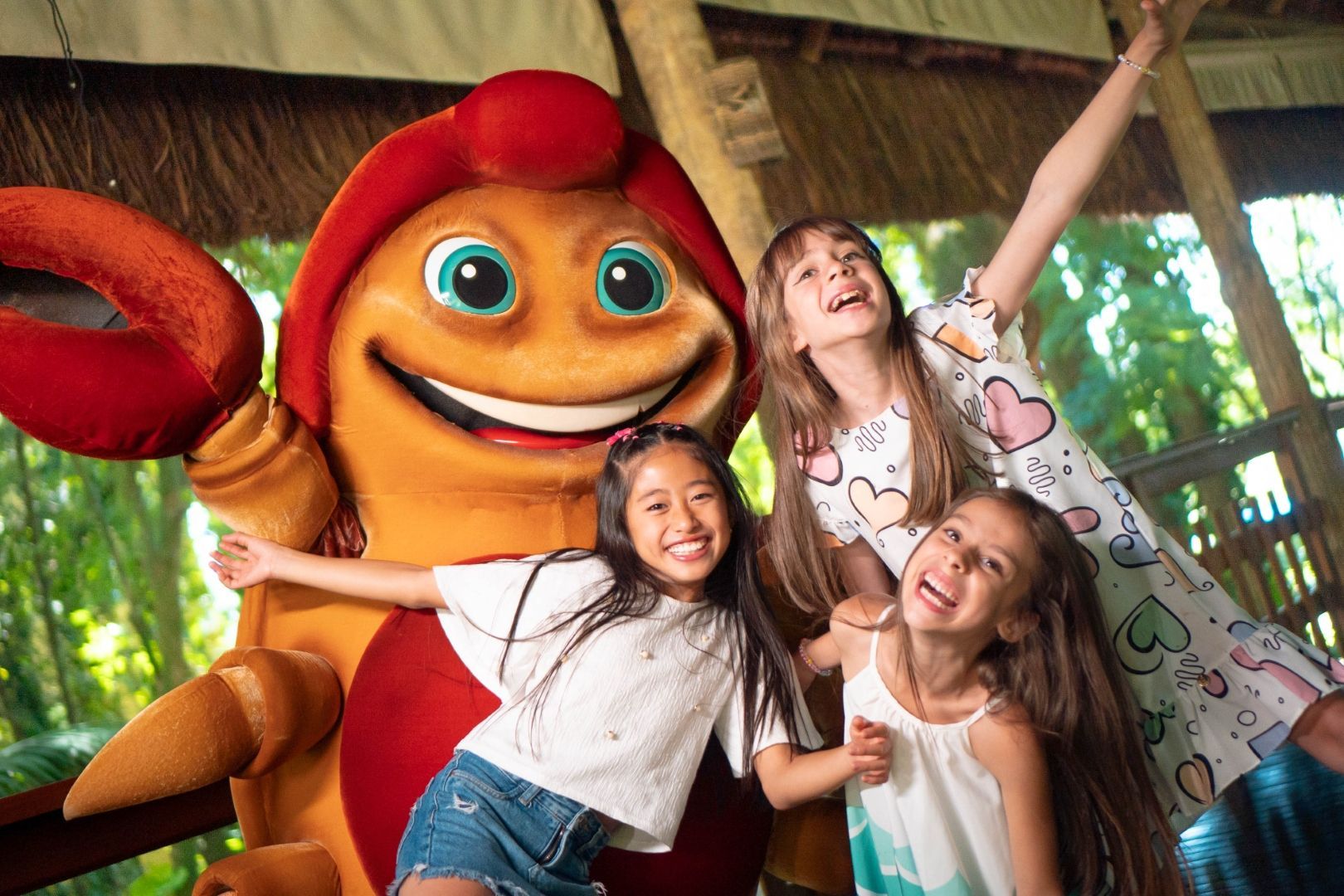 Três crianças posando com um grande mascote sorridente vermelho e laranja sob um telhado de palha.