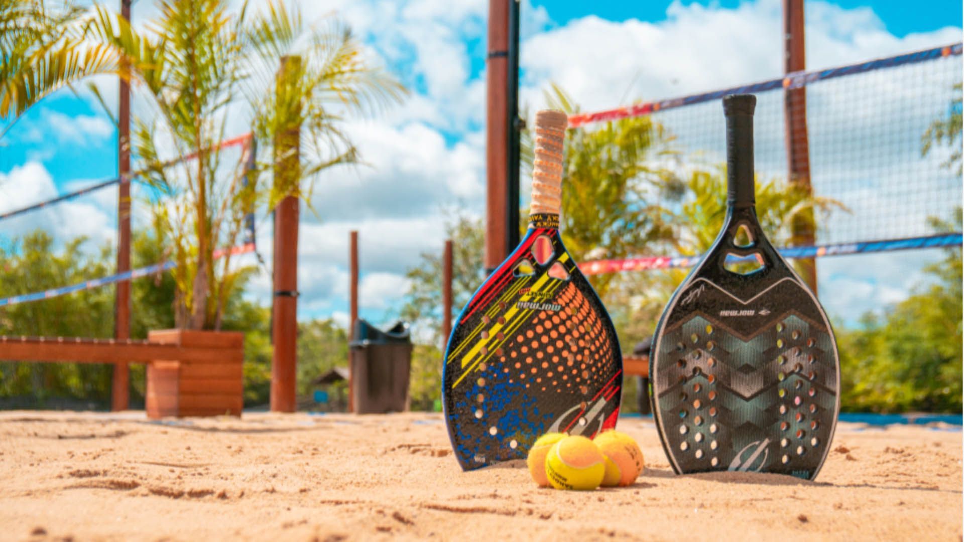 Duas raquetes de pickleball e uma bola em uma quadra de areia perto de uma rede, com palmeiras e céu azul ao fundo.