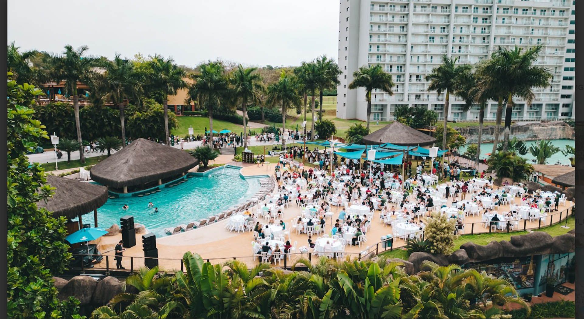 Piscina lotada de um resort com palmeiras, cabana e um grande hotel ao fundo.