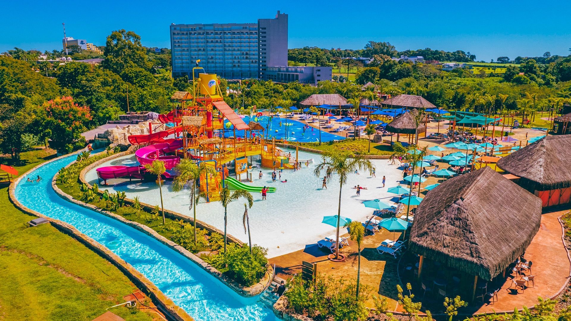 Parque aquático colorido com rio lento sinuoso, guarda-sóis, toboáguas e edifícios de resort sob um céu ensolarado.