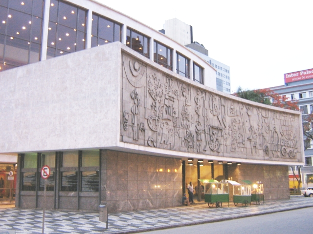 Fachada de edifício moderno com grande painel escultural em relevo e janelas no térreo, em uma rua da cidade.