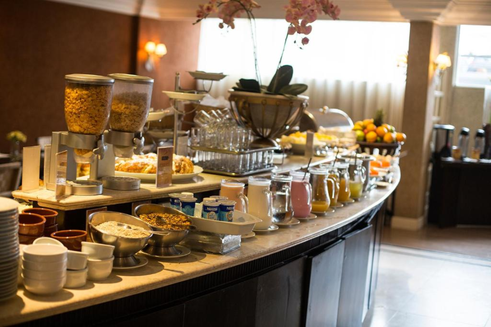 Buffet de café da manhã com doces, frutas, cereais, café e pratos empilhados em um balcão curvo.