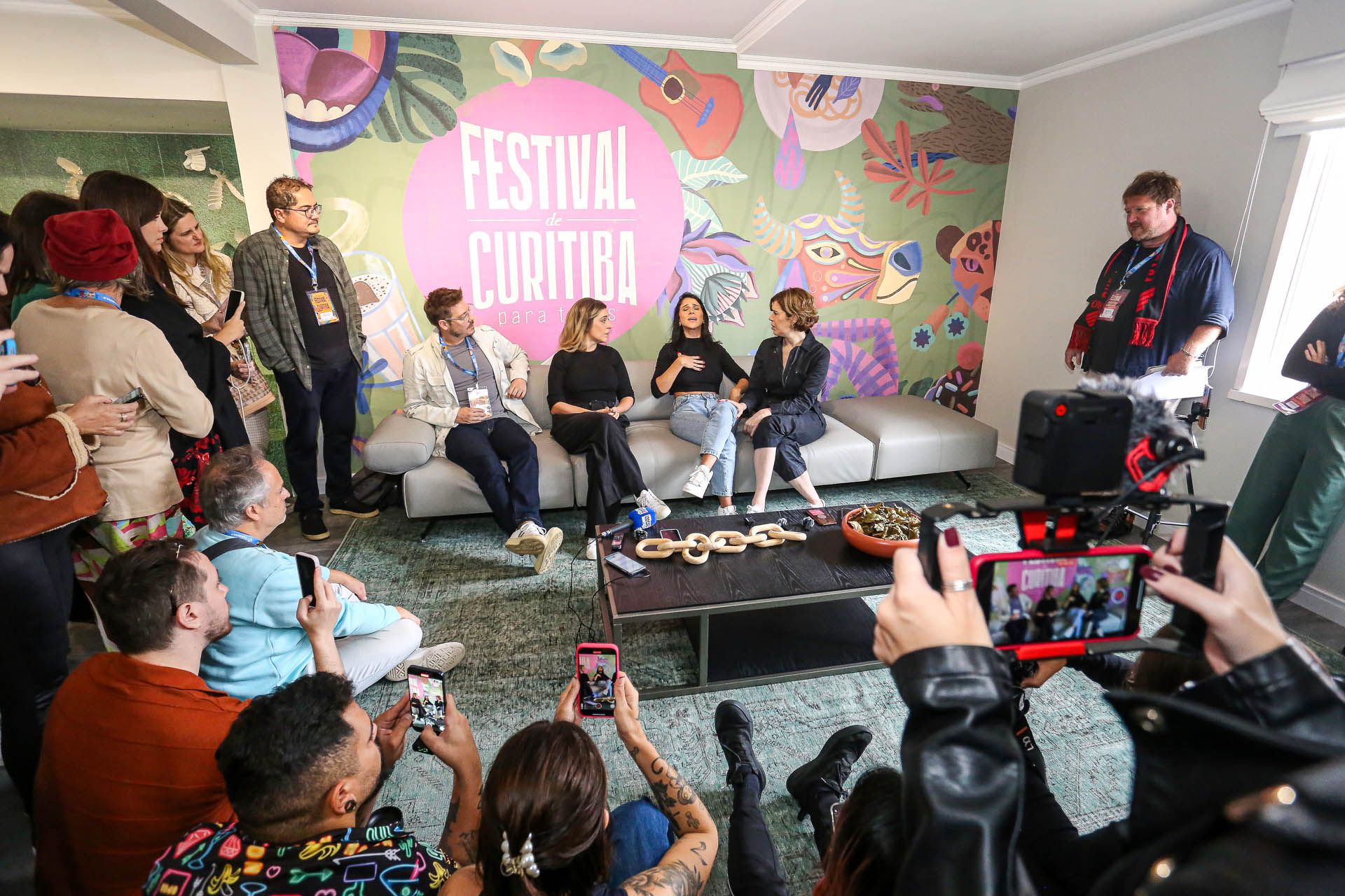 Mesa redonda em um evento de festival, com os palestrantes sentados em frente a um mural colorido e membros da plateia filmando.