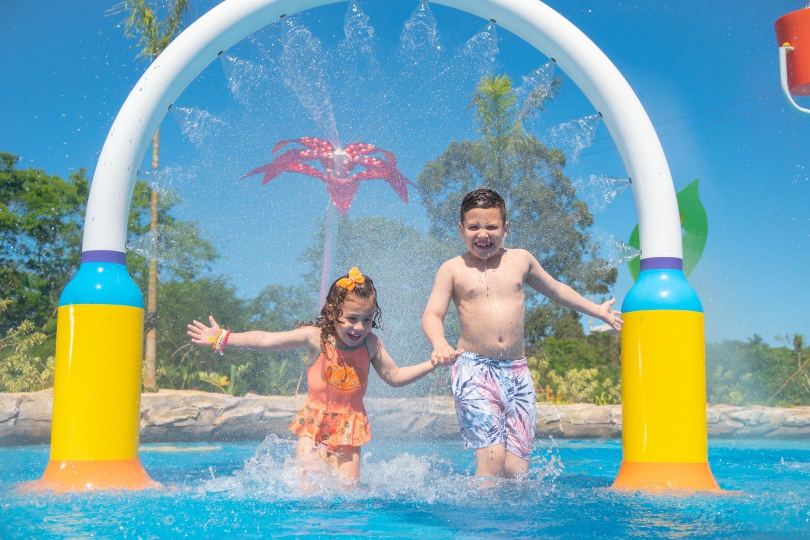 Duas crianças brincam na água sob um arco colorido em uma piscina ensolarada com palmeiras ao fundo.