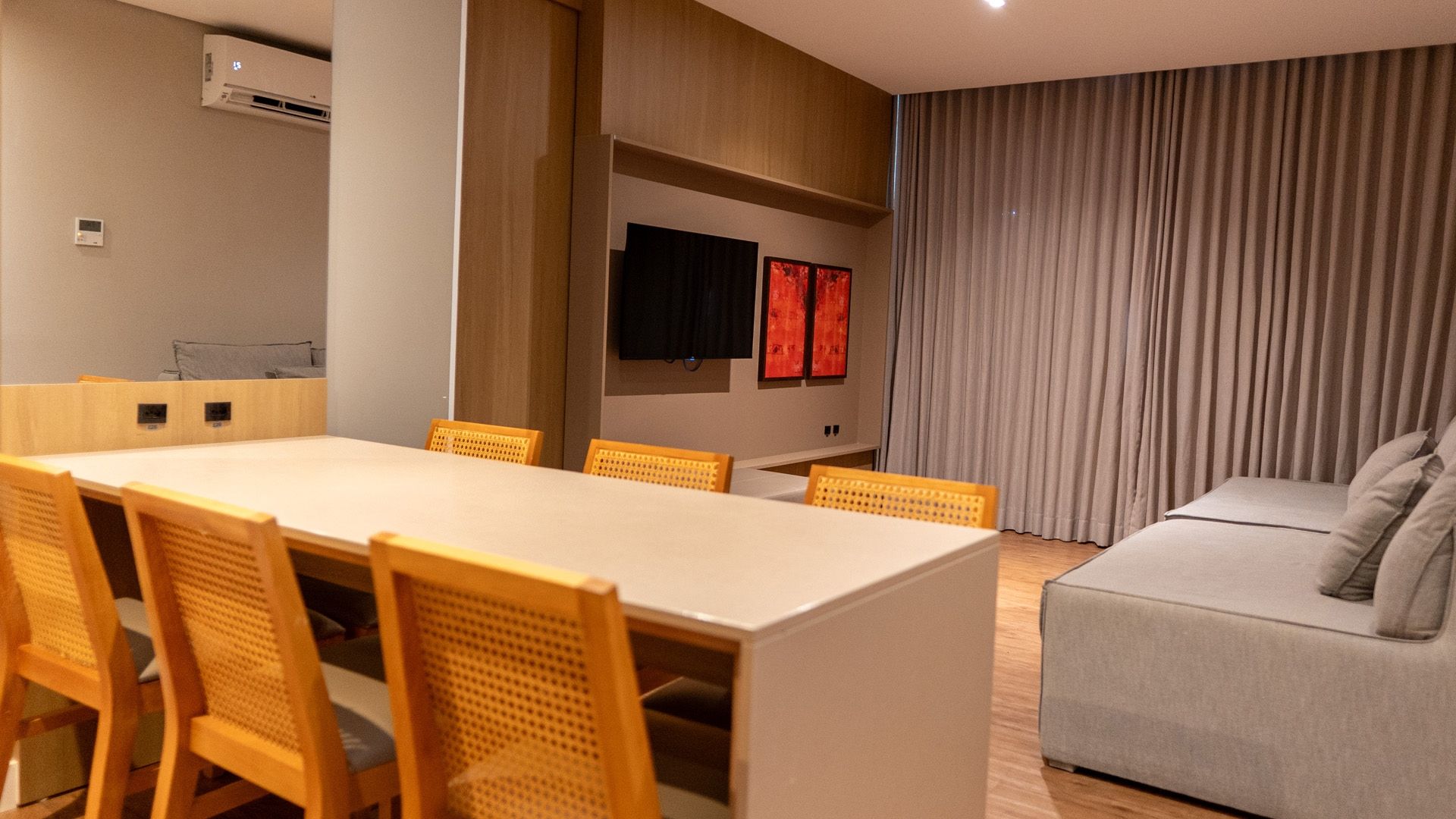 Sala de estar moderna com mesa de jantar, cadeiras laranja, TV de parede e sofá cinza junto às cortinas.