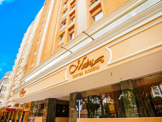 Exterior de um alto prédio de hotel bege com uma placa dourada e grandes janelas sob um céu azul brilhante.