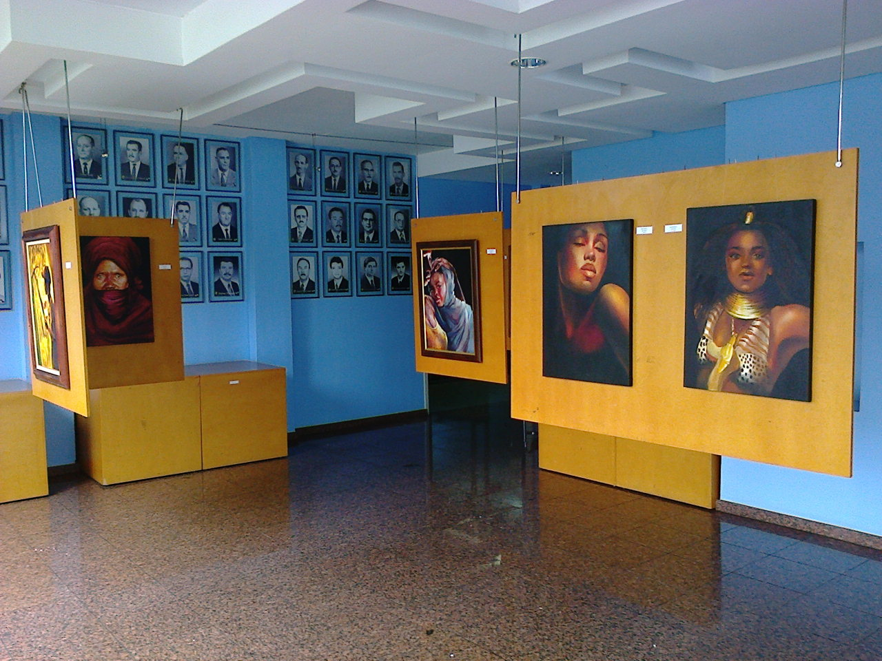 Galeria azul com retratos emoldurados em amarelo, expostos nas paredes e em painéis suspensos.