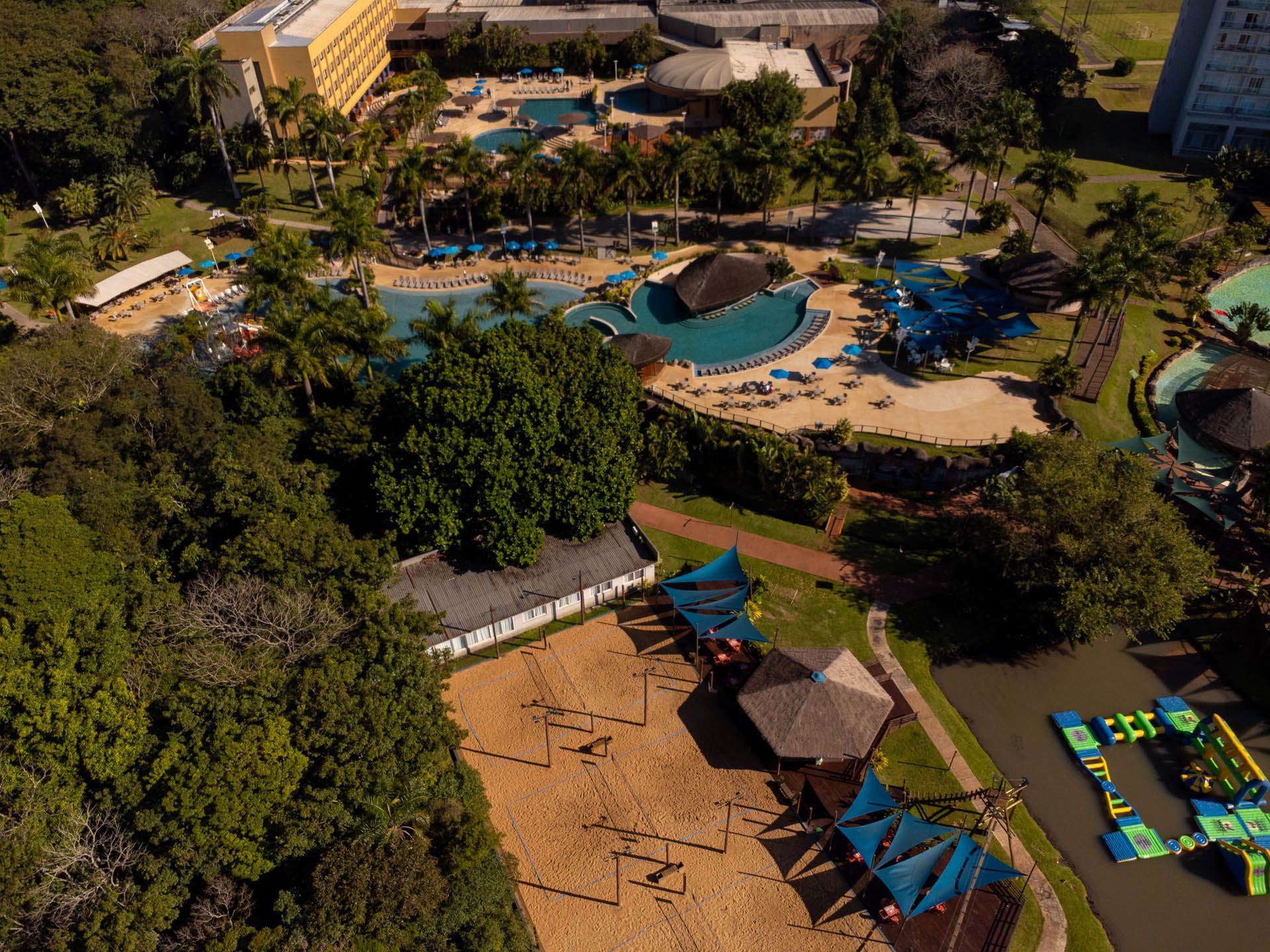 Vista aérea de um resort tropical com piscina, palmeiras e uma área de praia de areia com guarda-sóis azuis.