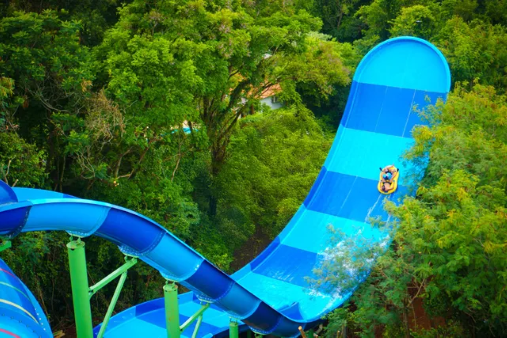 Pessoa deslizando por um toboágua azul em uma floresta verdejante.