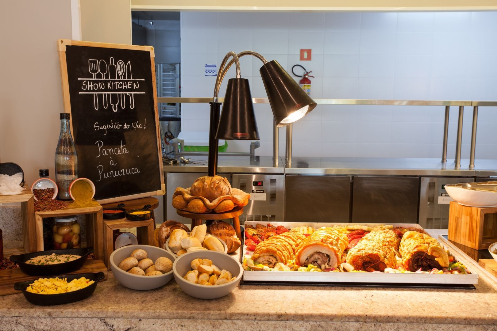Buffet de café da manhã com doces, frutas, muffins, pães e cardápio escrito em um quadro-negro sob luzes aconchegantes.