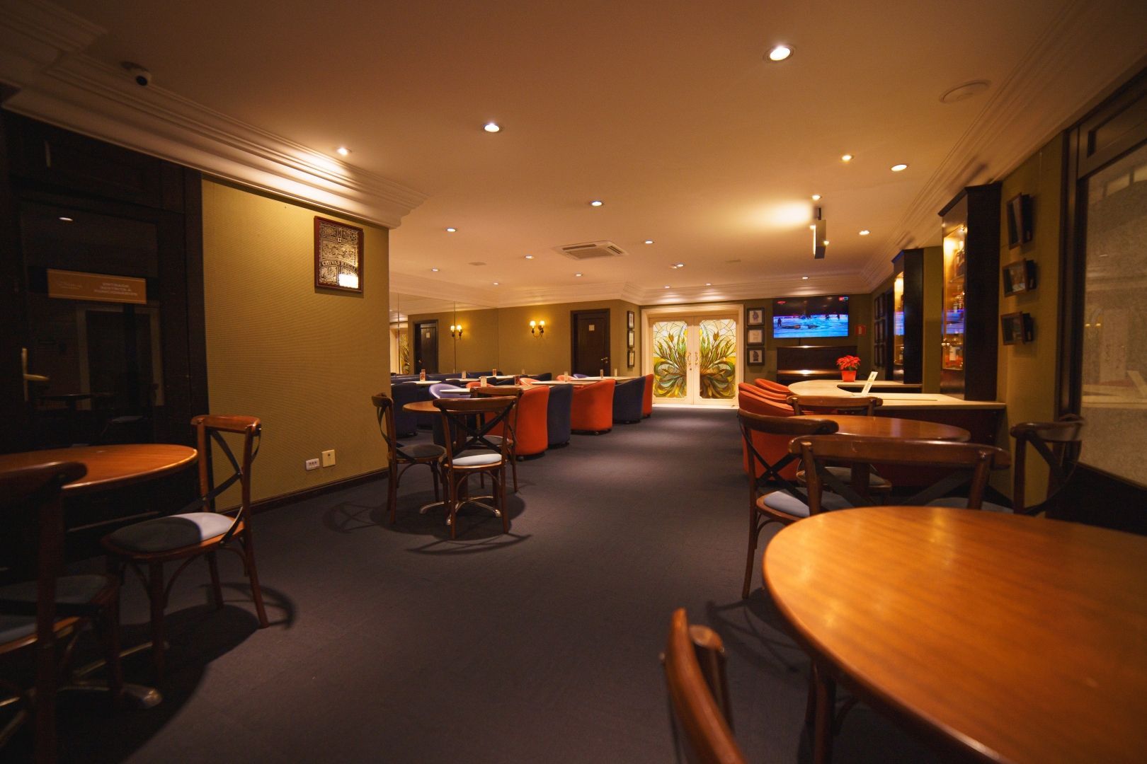 Salão de hotel vazio com mesas, cadeiras, iluminação aconchegante e um bar ao fundo.