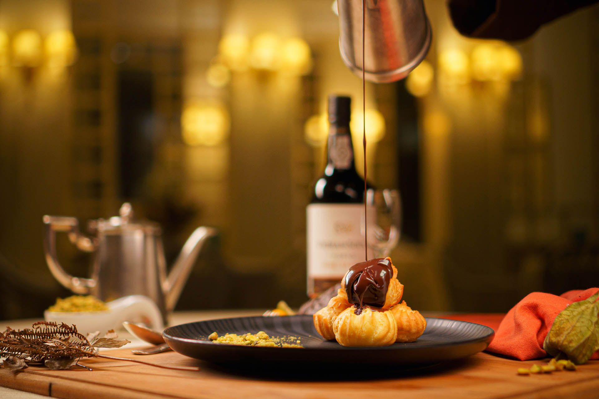 Sobremesa empratada com calda sendo regada, disposta sobre uma mesa de madeira com um bule de chá e uma garrafa de vinho sob luz quente.