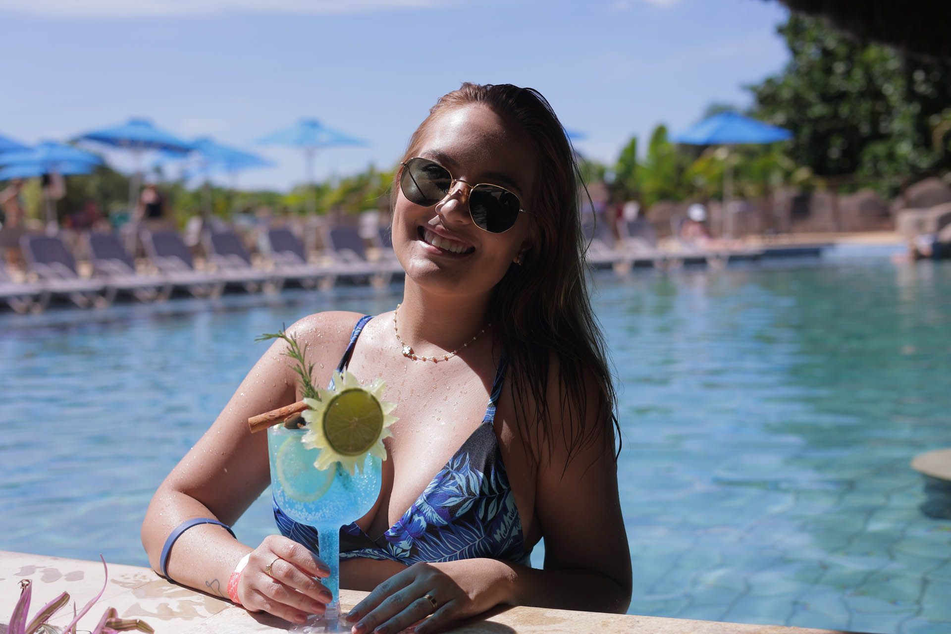 Mulher de óculos escuros segurando um coquetel azul à beira da piscina de um resort em um dia ensolarado.