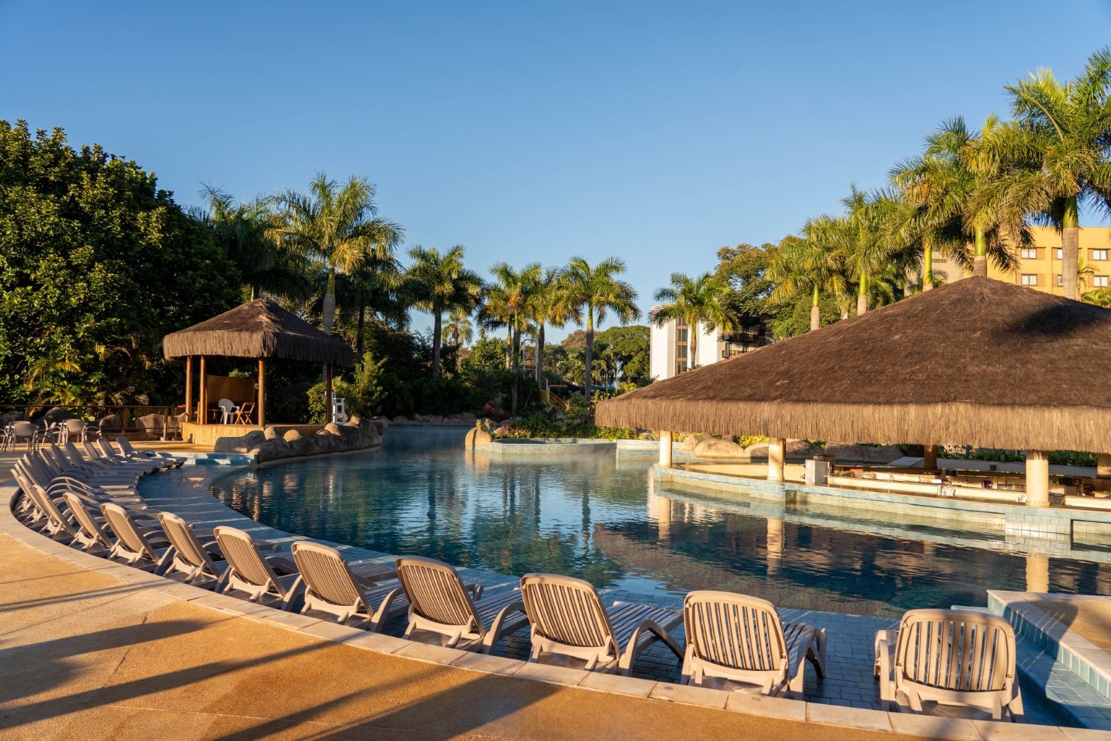 Resort à beira da piscina com espreguiçadeiras, palmeiras e cabanas de palha junto a uma lagoa tranquila ao pôr do sol.