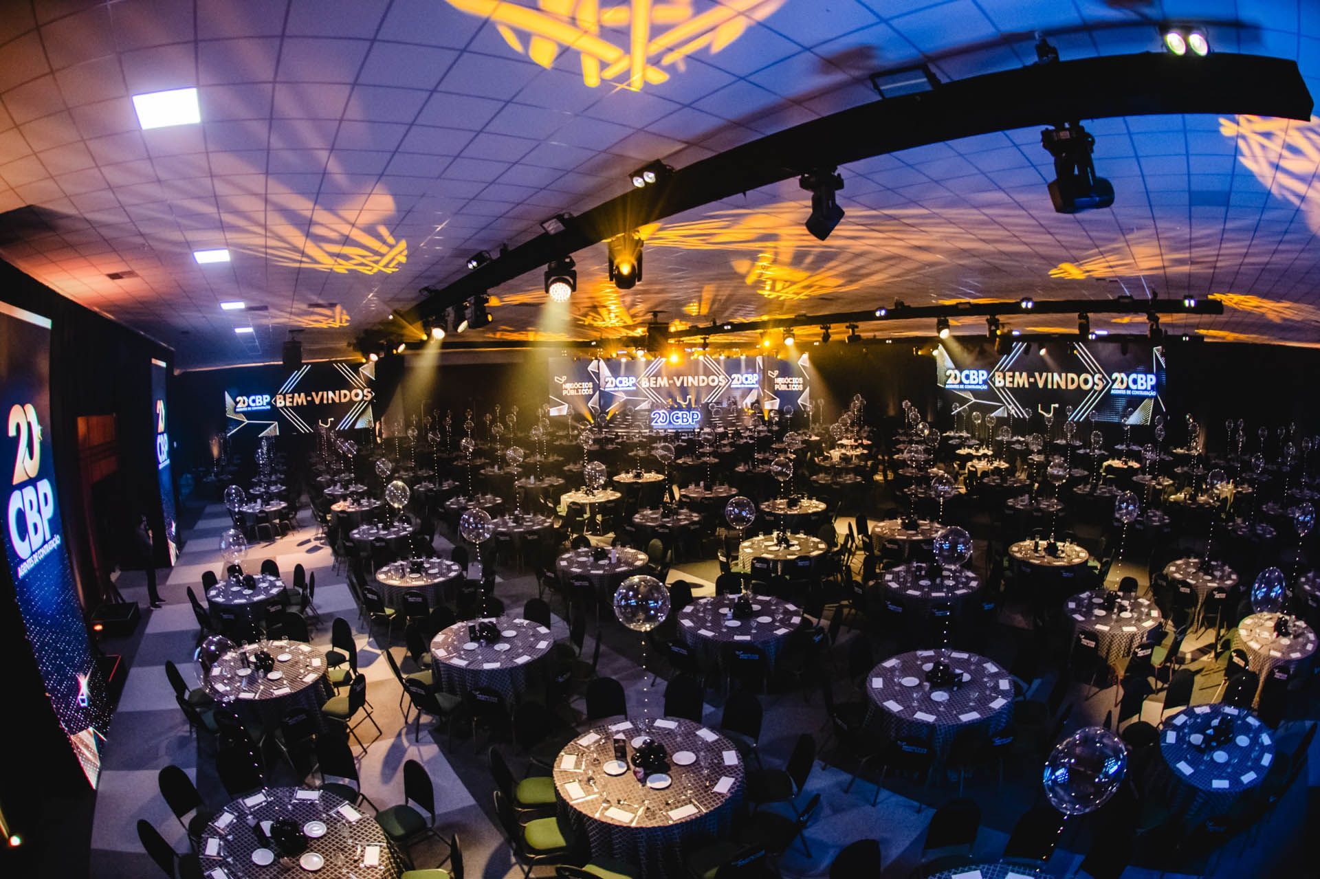 Amplo salão de banquetes com mesas redondas, iluminação azul-arroxeada e teto decorado antes de um evento.