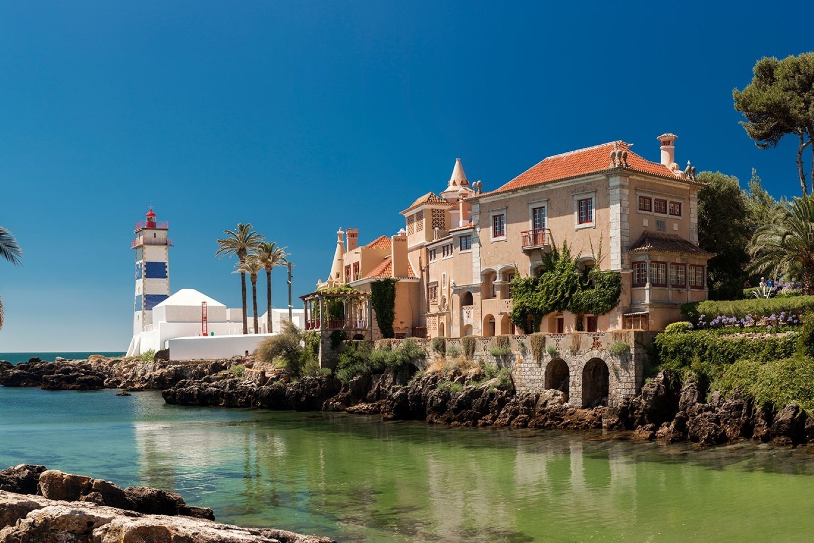Vila costeira com farol, construções de pedra e águas calmas e turquesas sob um céu azul brilhante.