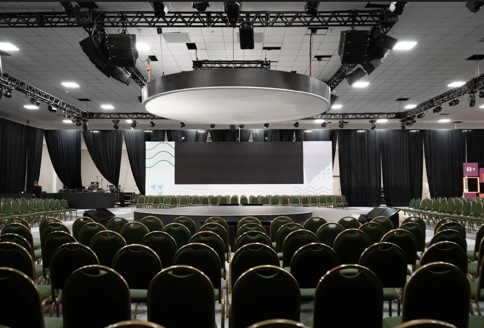 Sala de conferências vazia com fileiras de cadeiras verdes voltadas para um grande palco e tela de projeção.