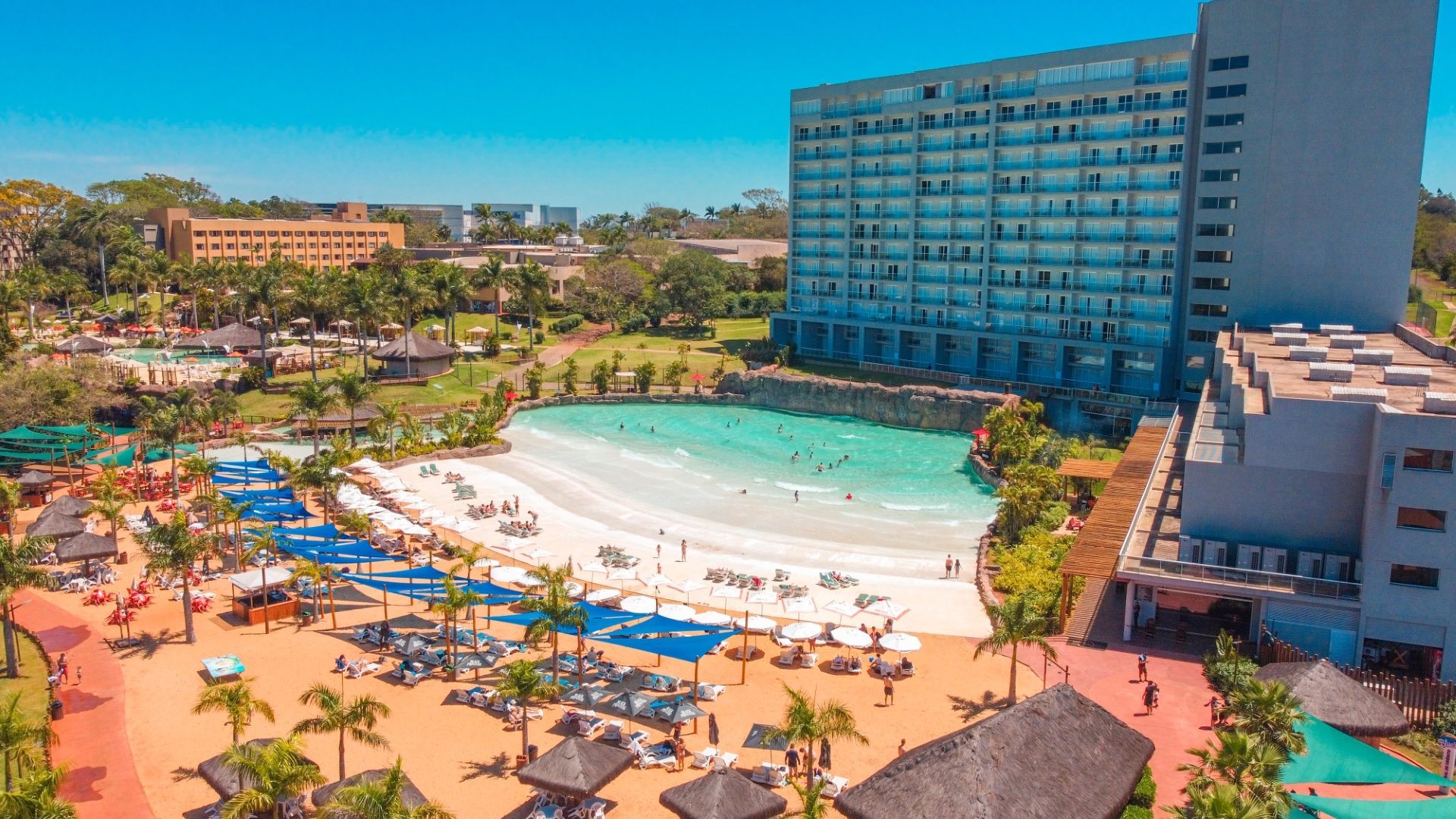 Piscina e área de praia do resort com água azul, espreguiçadeiras, guarda-sóis e um grande hotel com vista para a enseada.