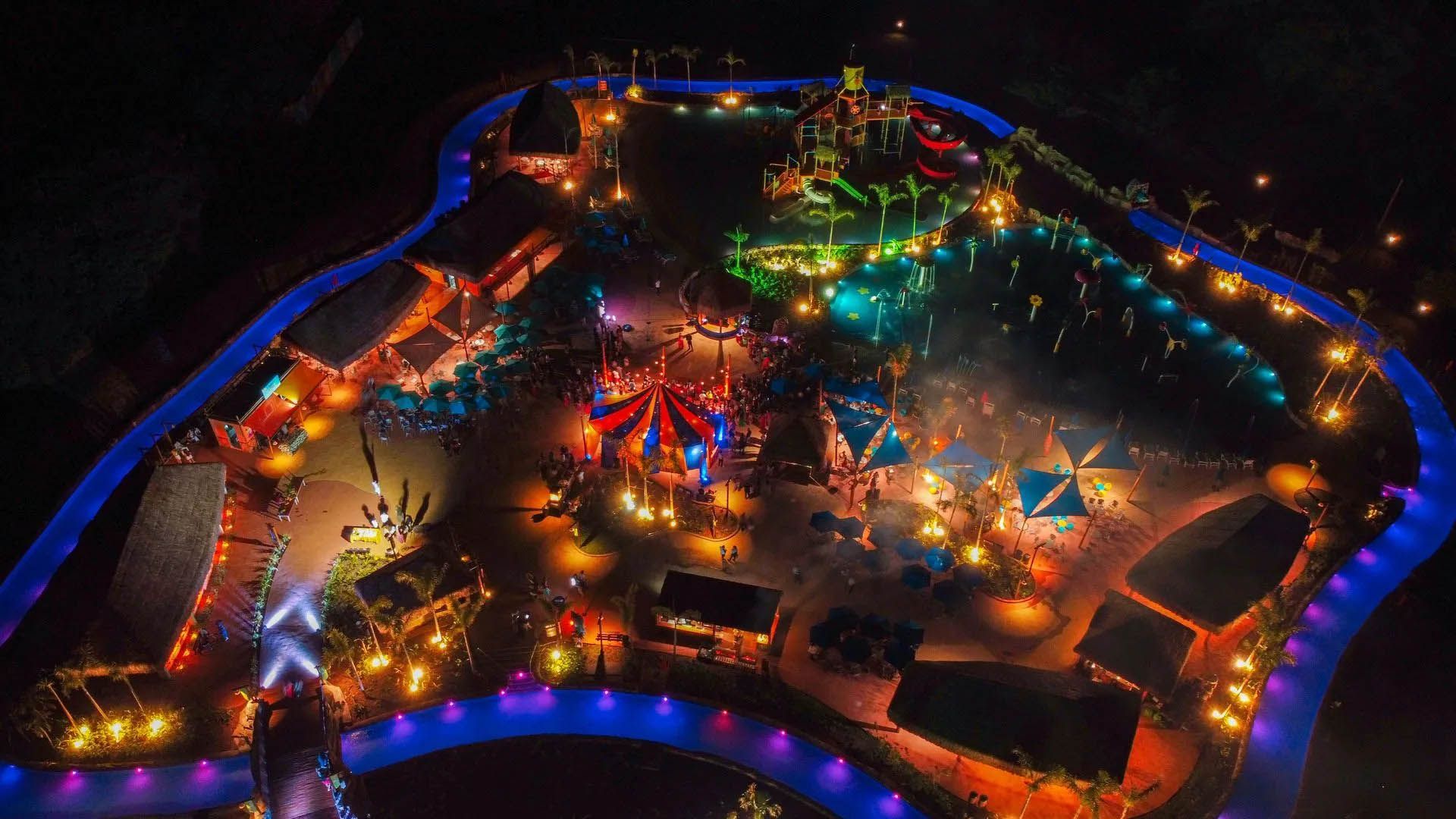 Vista aérea de um parque de diversões iluminado à noite, com brinquedos coloridos e luzes de néon.