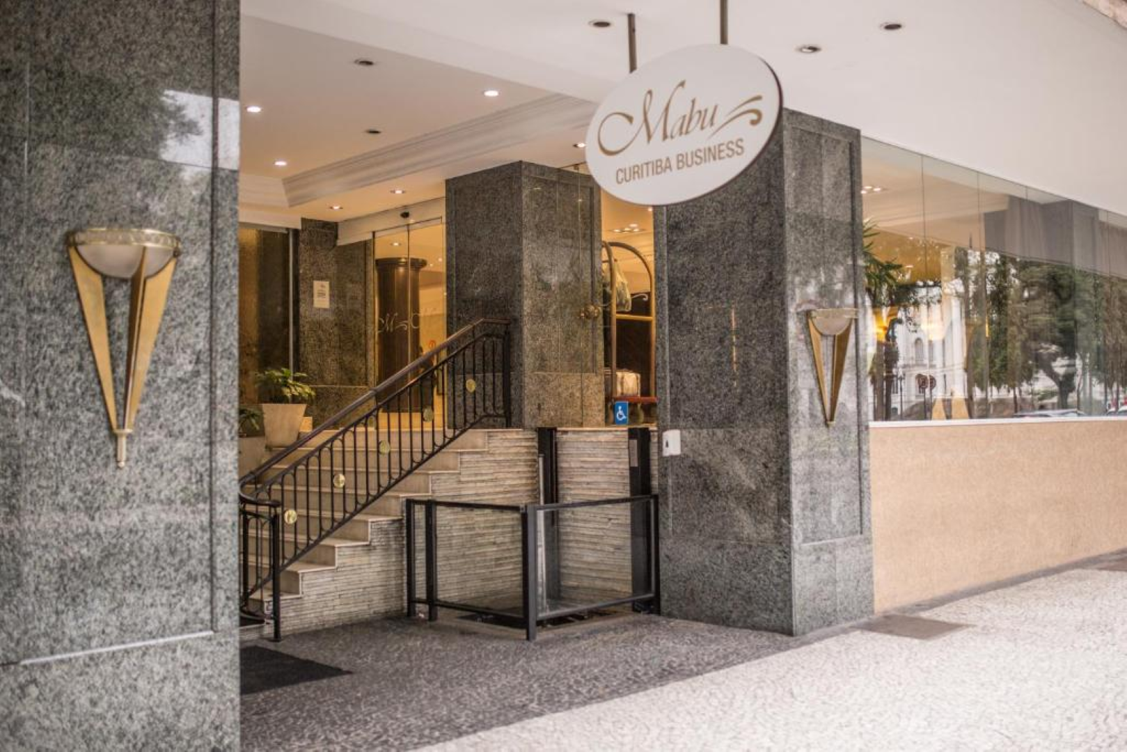 Entrada do edifício com colunas de mármore, escadas, portas de vidro e uma placa oval suspensa com a inscrição “Shiva”.